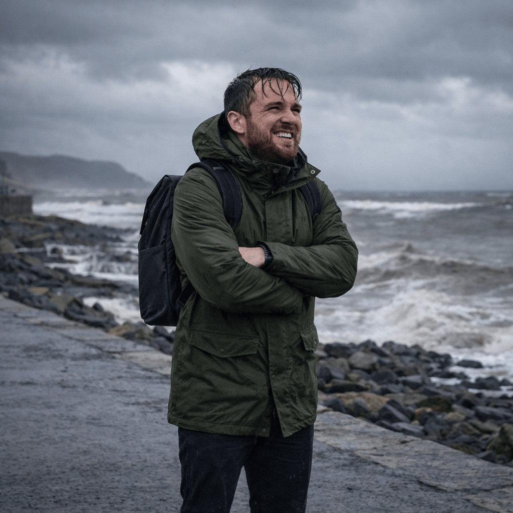 Man in green raincoat with backpack bracing against cold wind and rain by stormy seaside